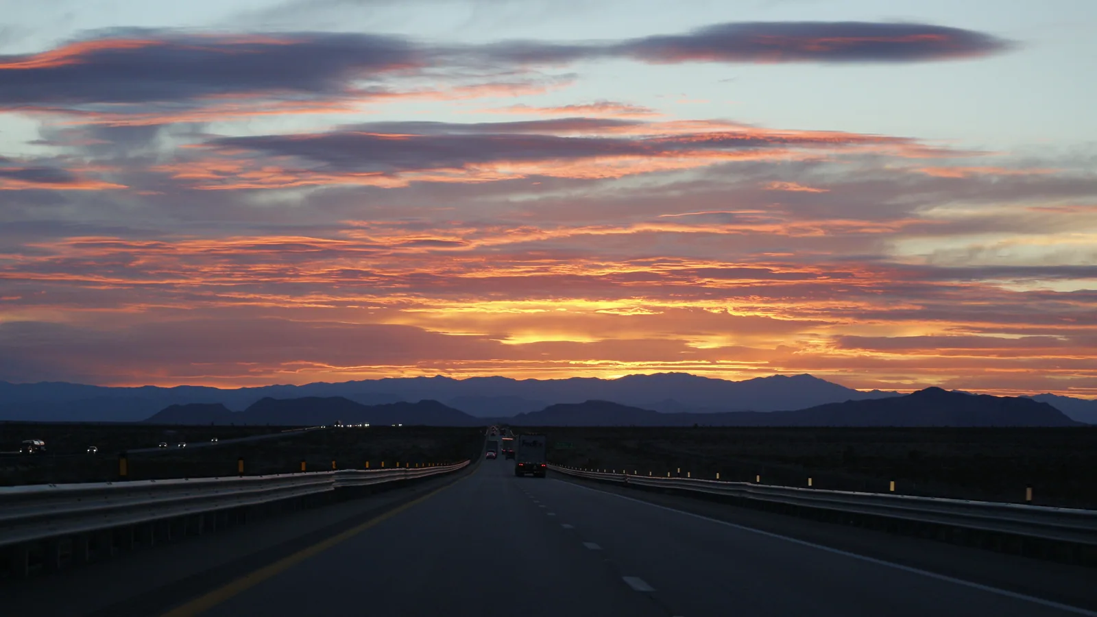 Interstate 15 near Glendale, Nevada at sunset, with a truck heading toward dark desert mountains under orange clouds