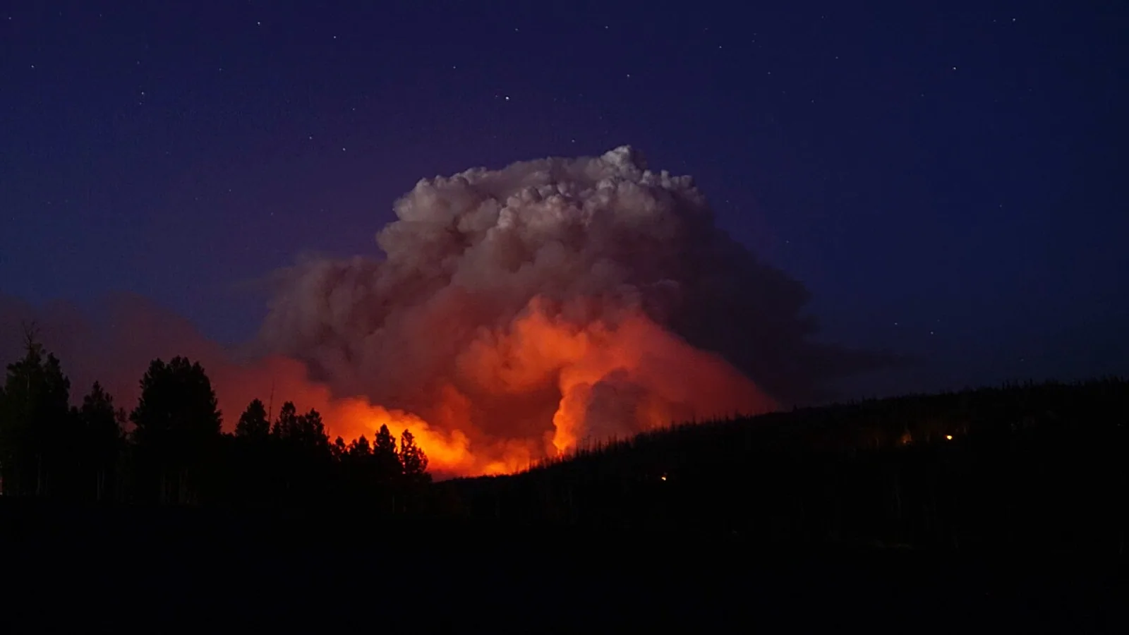A towering smoke column lit orange from below rises over a dark forested ridge under a starry night sky