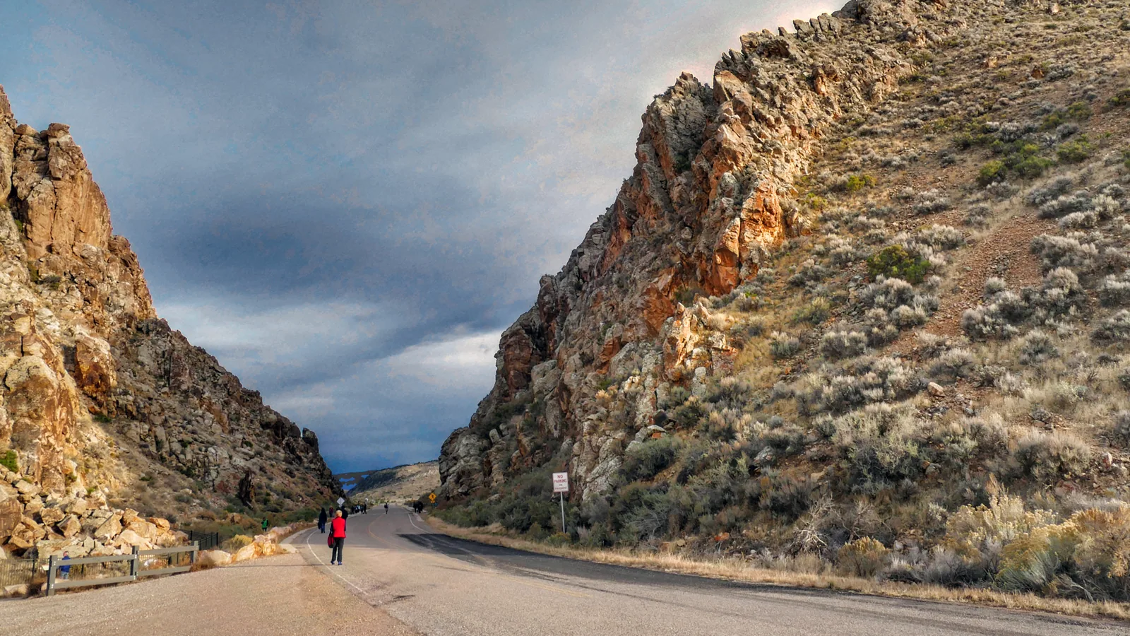 Gap Road die door de kloof van Parowan Gap loopt tussen rotsige heuvelruggen van de Red Hills onder een onweerslucht, met een paar mensen die langs de weg lopen