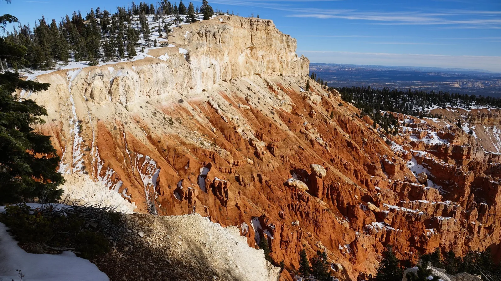 Snow on the orange and white cliffs below the Bristlecone Loop in Bryce Canyon National Park, with evergreens along the rim under a blue sky